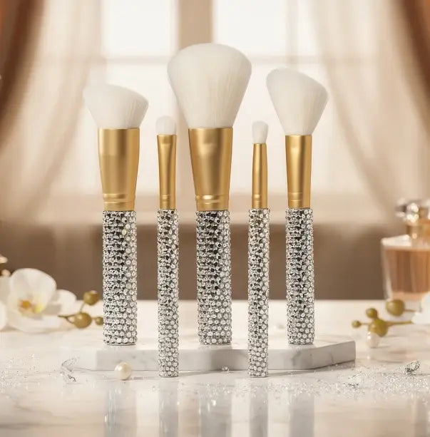 Rhinestone 5-Piece Makeup Brush Set by Genie displayed upright on a marble stand, showing crystal-encrusted handles, gold ferrules, and white bristles styled with white orchids and soft drapery background