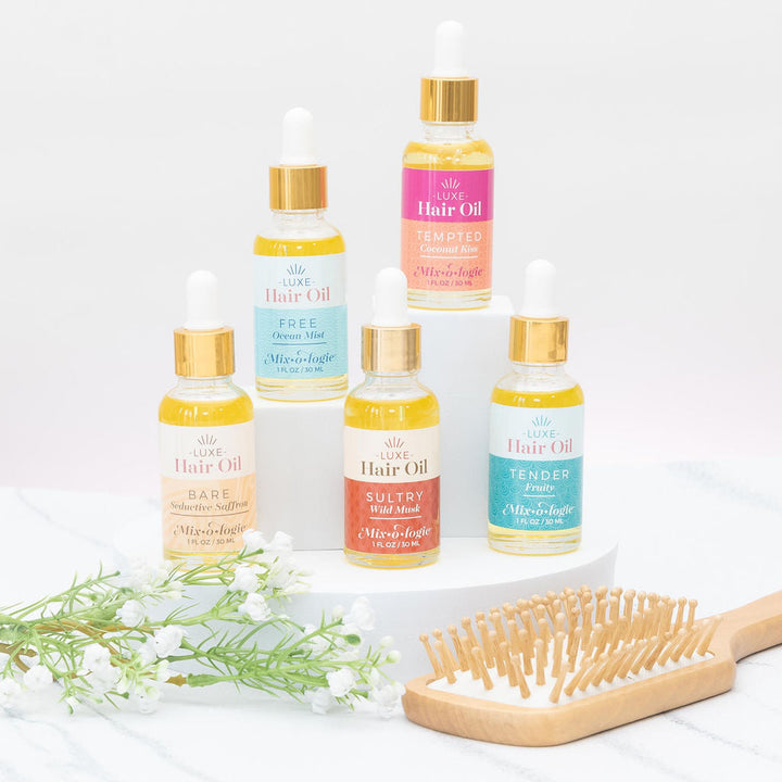 Mixologie Luxe Hair Oil collection, five scents displayed on white risers with a wood hairbrush and white florals — Bare, Free, Tempted, Sultry, and Tender
