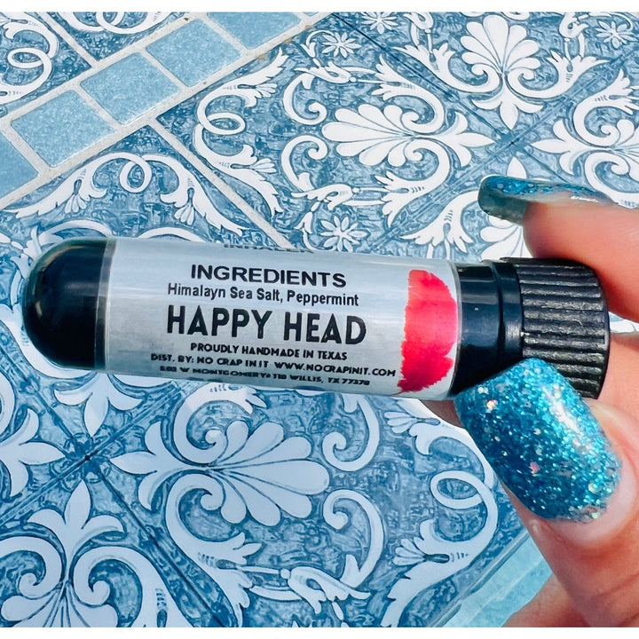 Hand with teal glitter nails holding a No Crap In It Happy Head essential inhaler with black cap, made with Himalayan sea salt and peppermint, on a blue decorative mosaic tile surface.