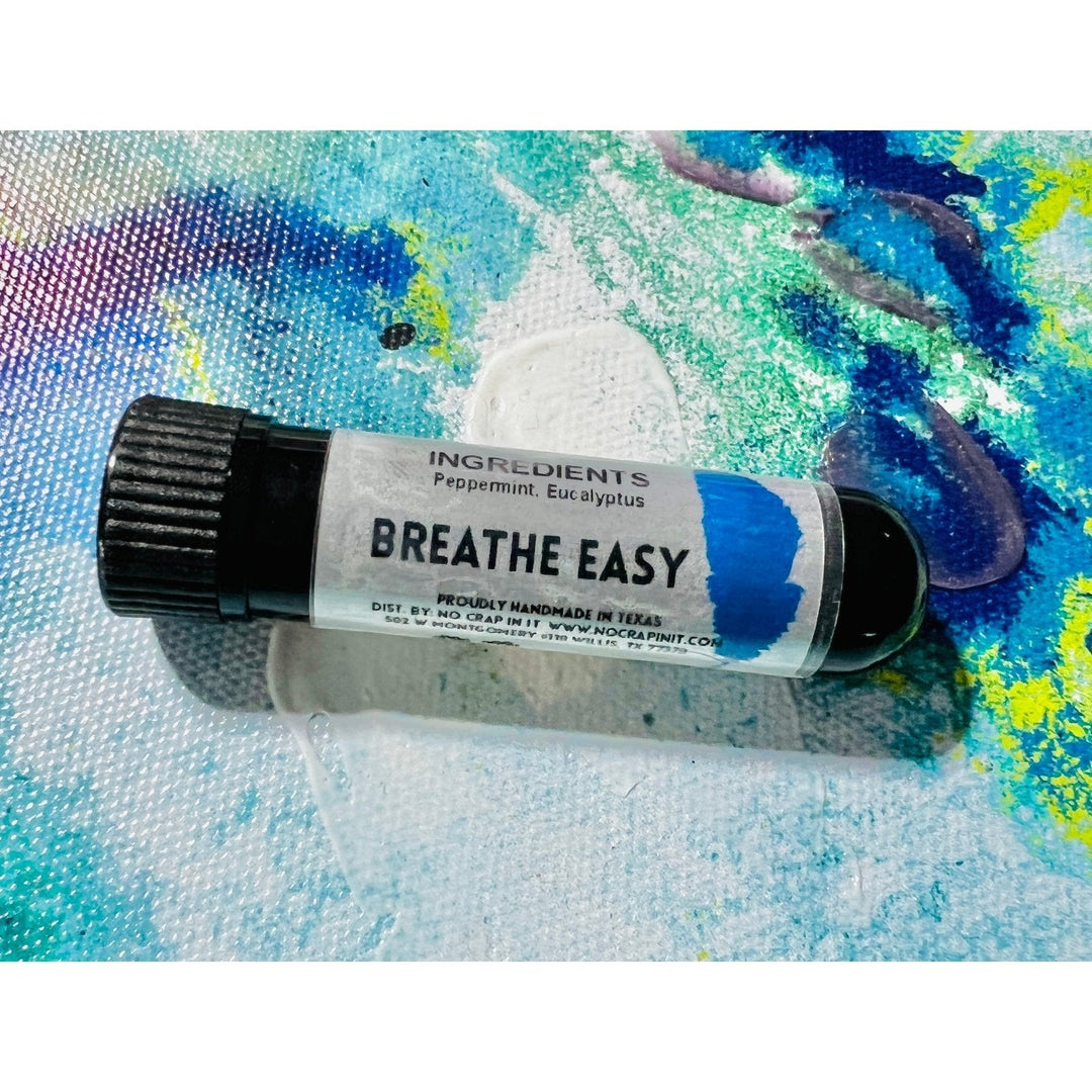 No Crap In It Breathe Easy essential inhaler with black cap, made with peppermint and eucalyptus, lying on a colorful blue, green, and yellow paint splatter background.