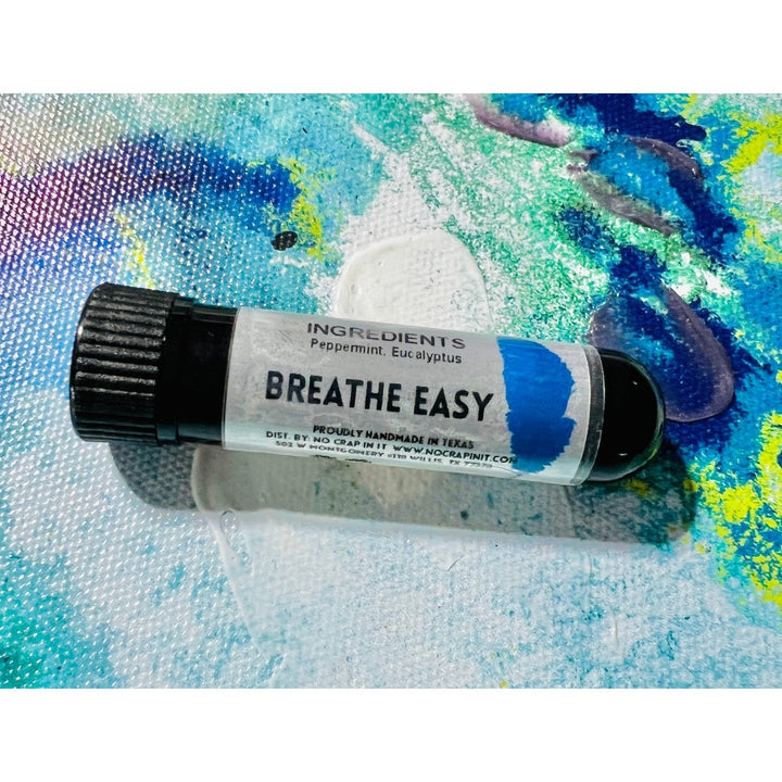 No Crap In It Breathe Easy essential inhaler with black cap, made with peppermint and eucalyptus, lying on a colorful blue, green, and yellow paint splatter background.