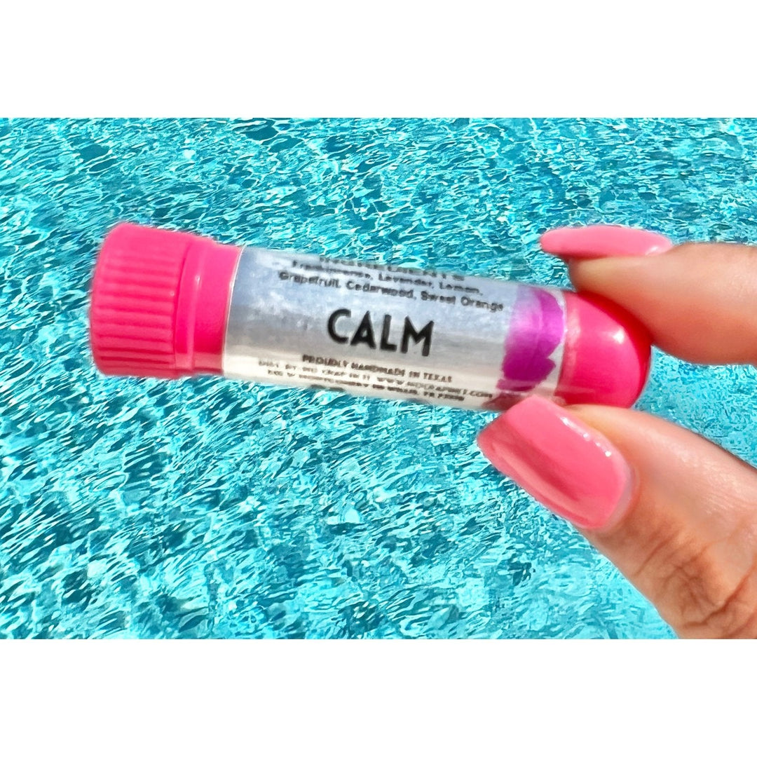 Hand with pink nails holding a No Crap In It Calm essential inhaler with hot pink cap, made with frankincense, lavender, lemon, grapefruit, cedarwood, and sweet orange, over a bright blue swimming pool.