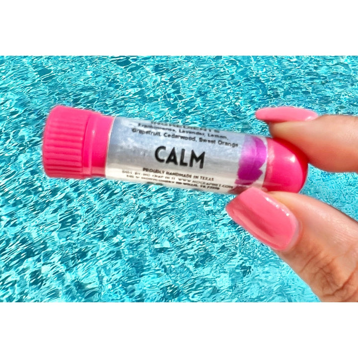 Hand with pink nails holding a No Crap In It Calm essential inhaler with hot pink cap, made with frankincense, lavender, lemon, grapefruit, cedarwood, and sweet orange, over a bright blue swimming pool.