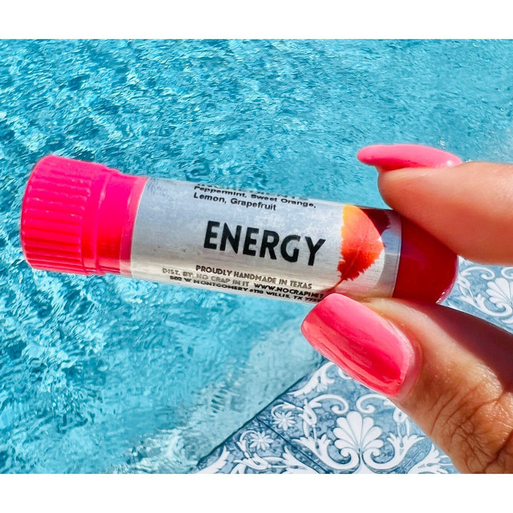 Hand with hot pink nails holding a No Crap In It Energy essential inhaler with hot pink cap, made with peppermint, sweet orange, lemon, and grapefruit, over a bright blue swimming pool.