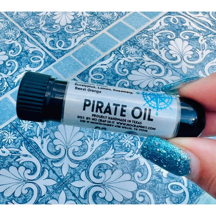 Hand with teal glitter nails holding a No Crap In It Pirate Oil essential inhaler with black cap, made with eucalyptus, lemon, rosemary, and sweet orange, on a blue decorative mosaic tile surface.