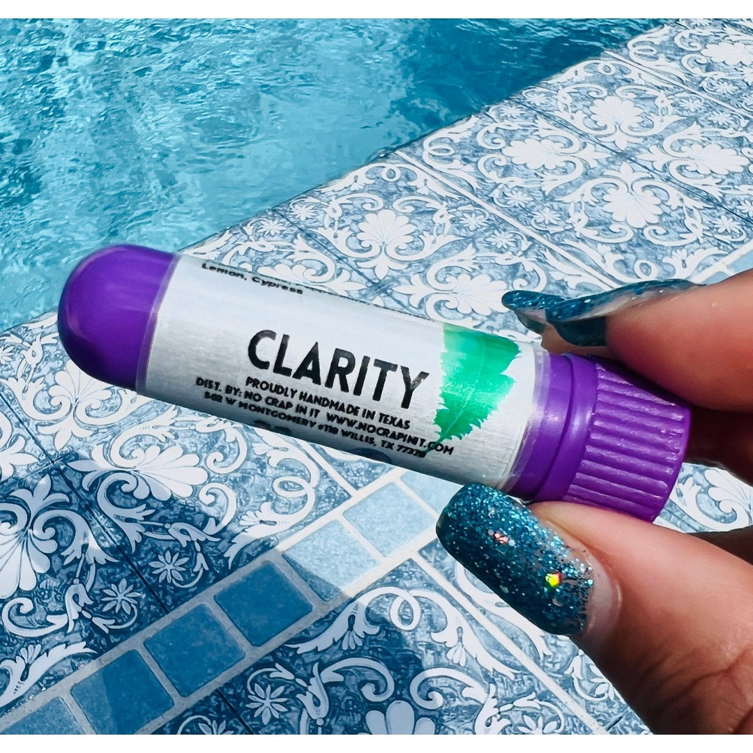 Hand with teal glitter nails holding a No Crap In It Clarity essential inhaler with purple cap, made with lemon and cypress, poolside on a blue mosaic tile surface.