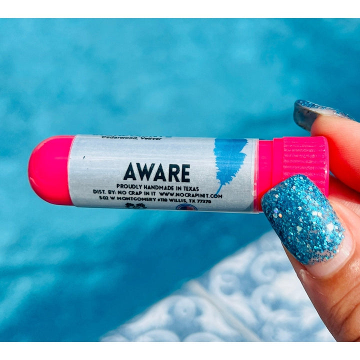 Hand with teal glitter nails holding a No Crap In It Aware essential inhaler with hot pink cap, made with cedarwood and vetiver, over a bright blue swimming pool.