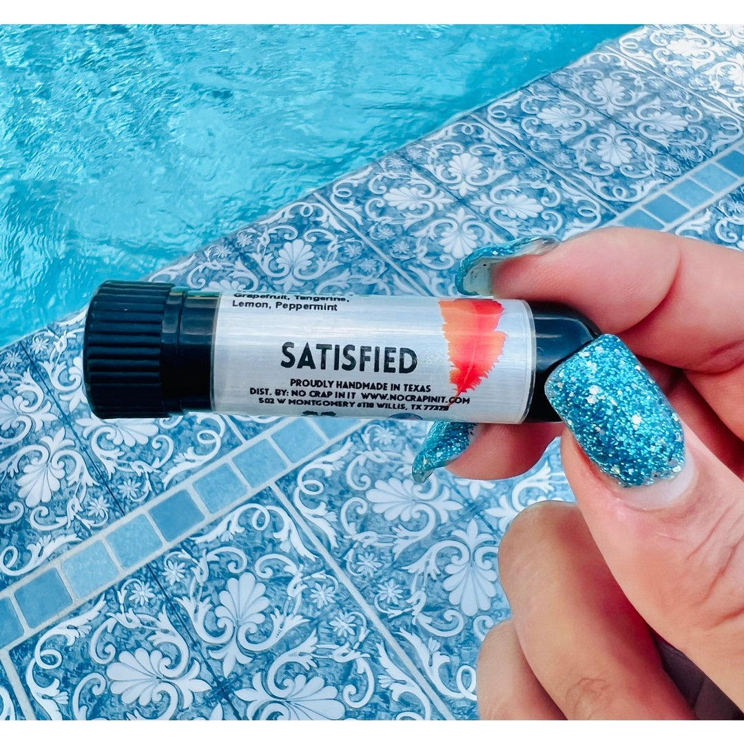 Hand with teal glitter nails holding a No Crap In It Satisfied essential inhaler with black cap, made with grapefruit, tangerine, lemon, and peppermint, poolside on a blue mosaic tile surface.