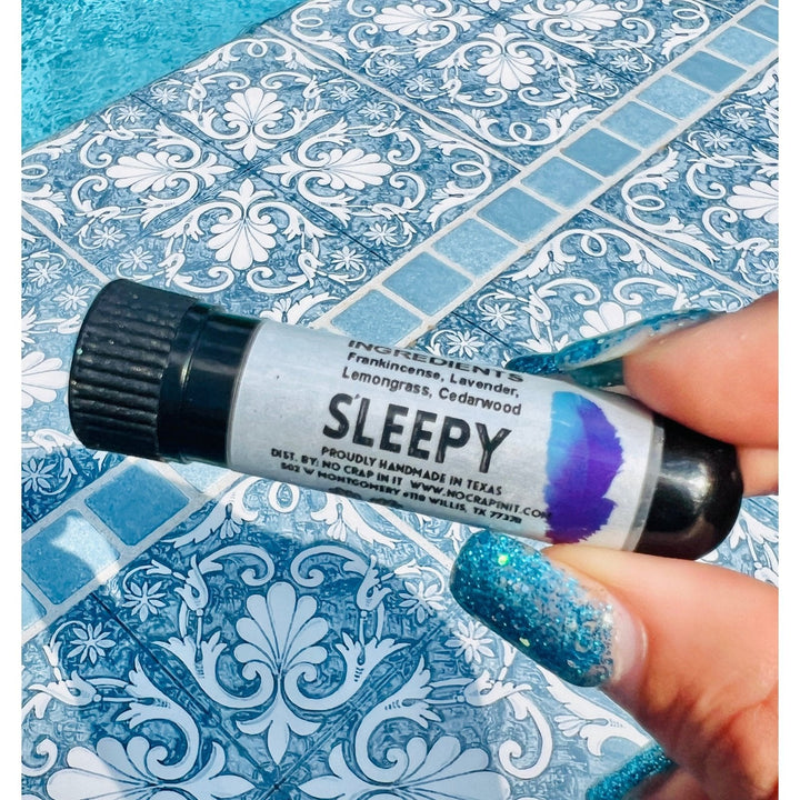 Hand with teal glitter nails holding a No Crap In It Sleepy essential inhaler with black cap, made with frankincense, lavender, lemongrass, and cedarwood, poolside on a blue mosaic tile surface.