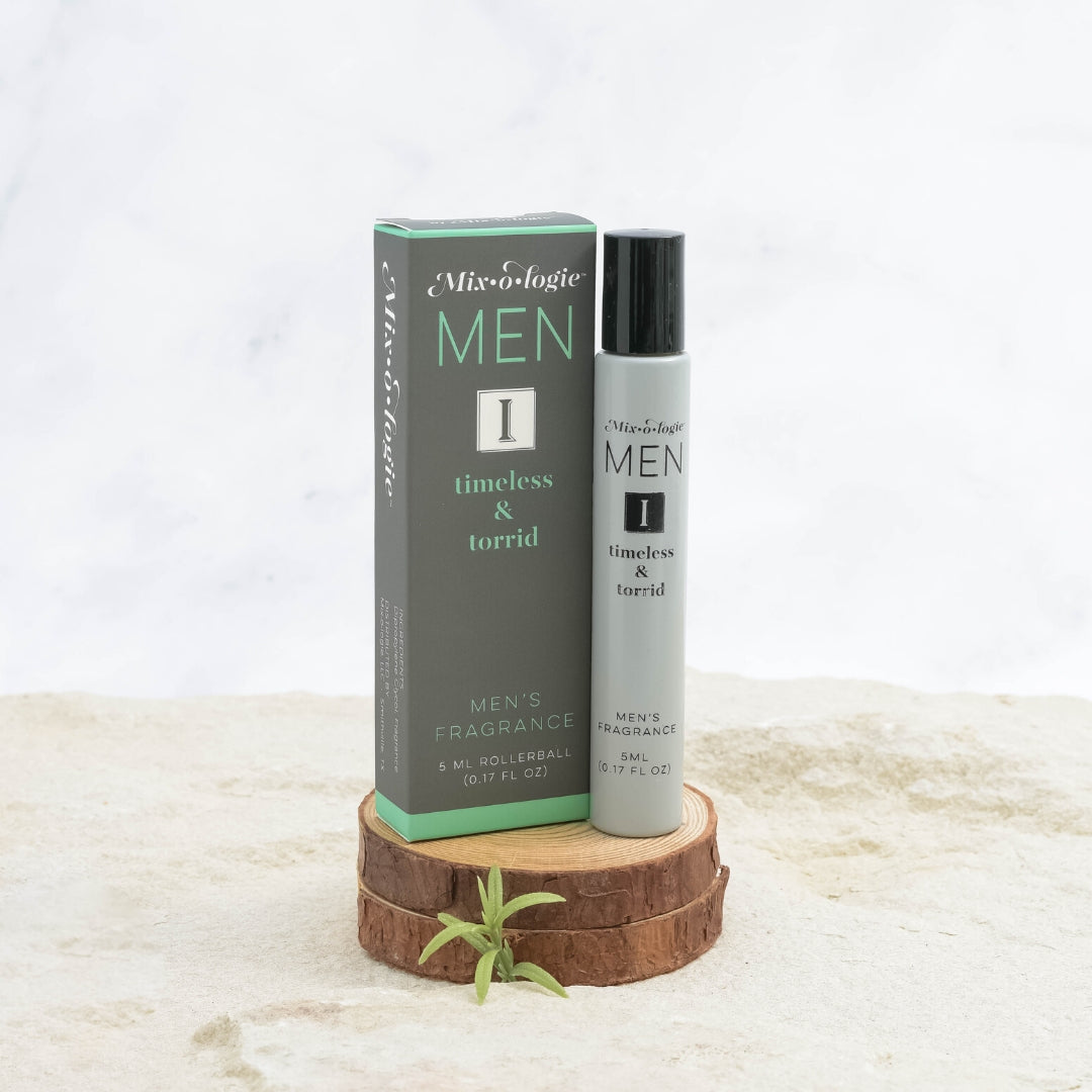 Mixologie Men's Fragrance I Timeless & Torrid, 5 ml rollerball, gray bottle with black cap next to dark gray and teal box displayed on stacked wood slices with small green sprig on marble surface