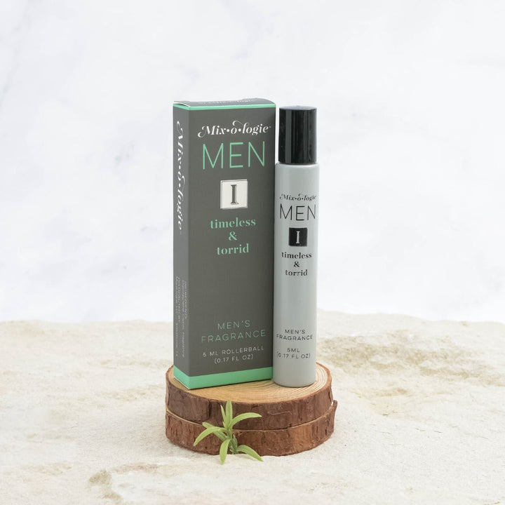 Mixologie Men's Fragrance I Timeless & Torrid, 5 ml rollerball, gray bottle with black cap next to dark gray and teal box displayed on stacked wood slices with small green sprig on marble surface