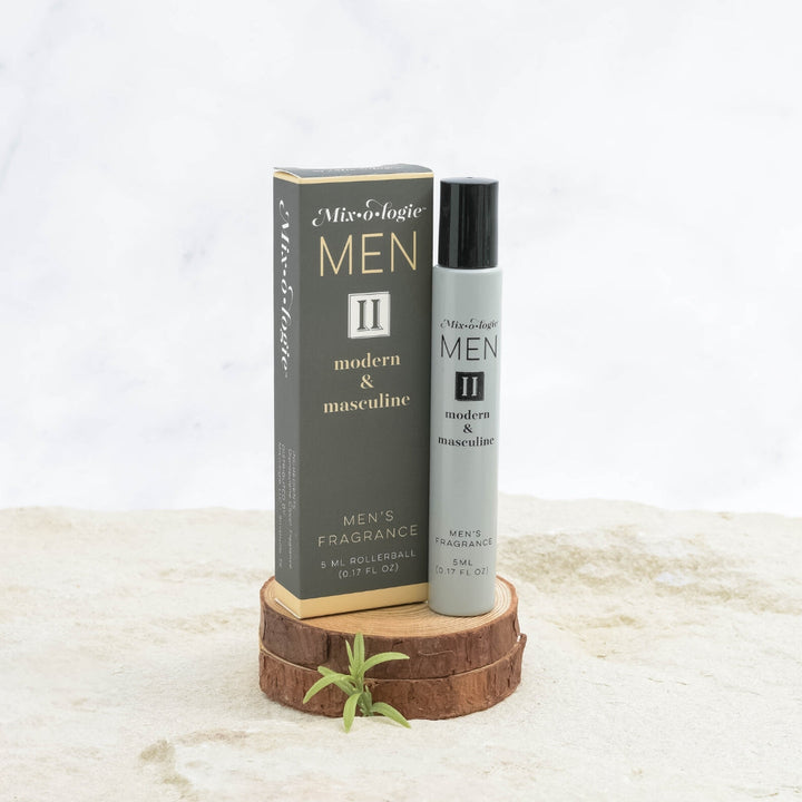 Mixologie Men's Fragrance II Modern & Masculine, 5 ml rollerball, gray bottle with black cap next to dark gray box with gold accent band displayed on stacked wood slices with small green sprig on marble surface