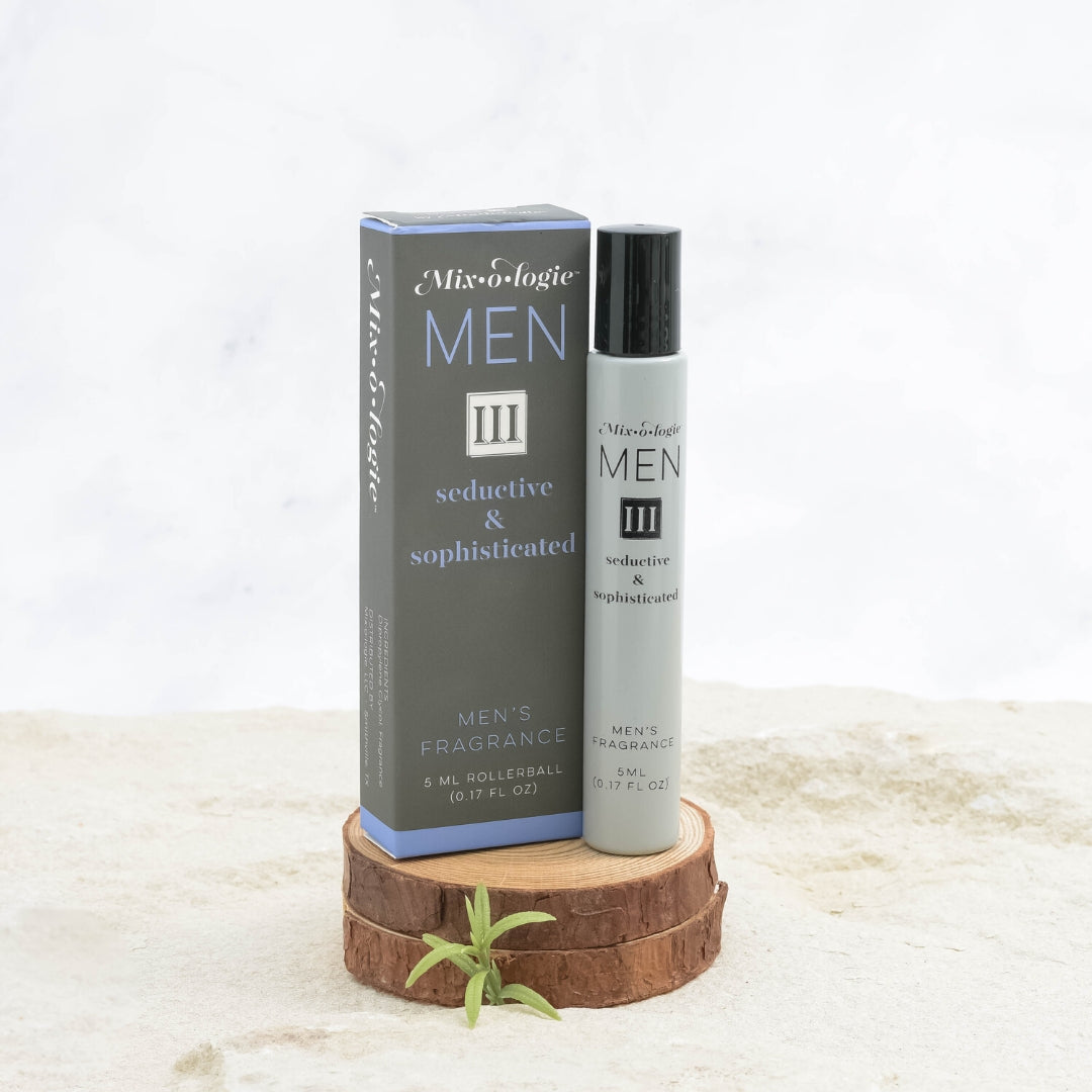 Mixologie Men's Fragrance III Seductive & Sophisticated, 5 ml rollerball, gray bottle with black cap next to dark gray and periwinkle blue box displayed on stacked wood slices with small green sprig on marble surface