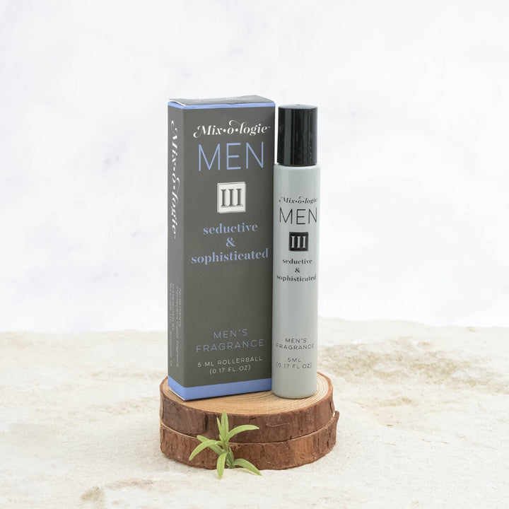 Mixologie Men's Fragrance III Seductive & Sophisticated, 5 ml rollerball, gray bottle with black cap next to dark gray and periwinkle blue box displayed on stacked wood slices with small green sprig on marble surface