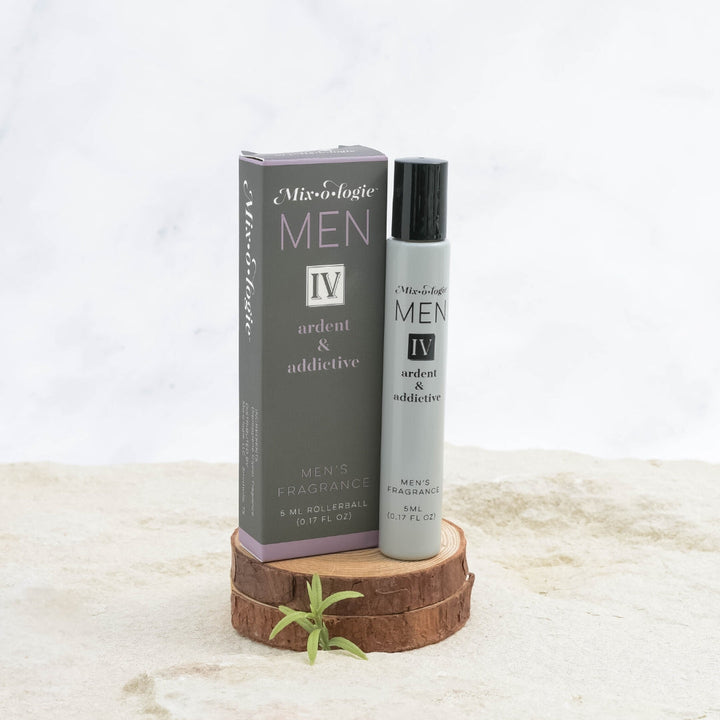 Mixologie Men's Fragrance IV Ardent & Addictive, 5 ml rollerball, gray bottle with black cap next to dark gray box with lavender accent band displayed on stacked wood slices with small green sprig on marble surface