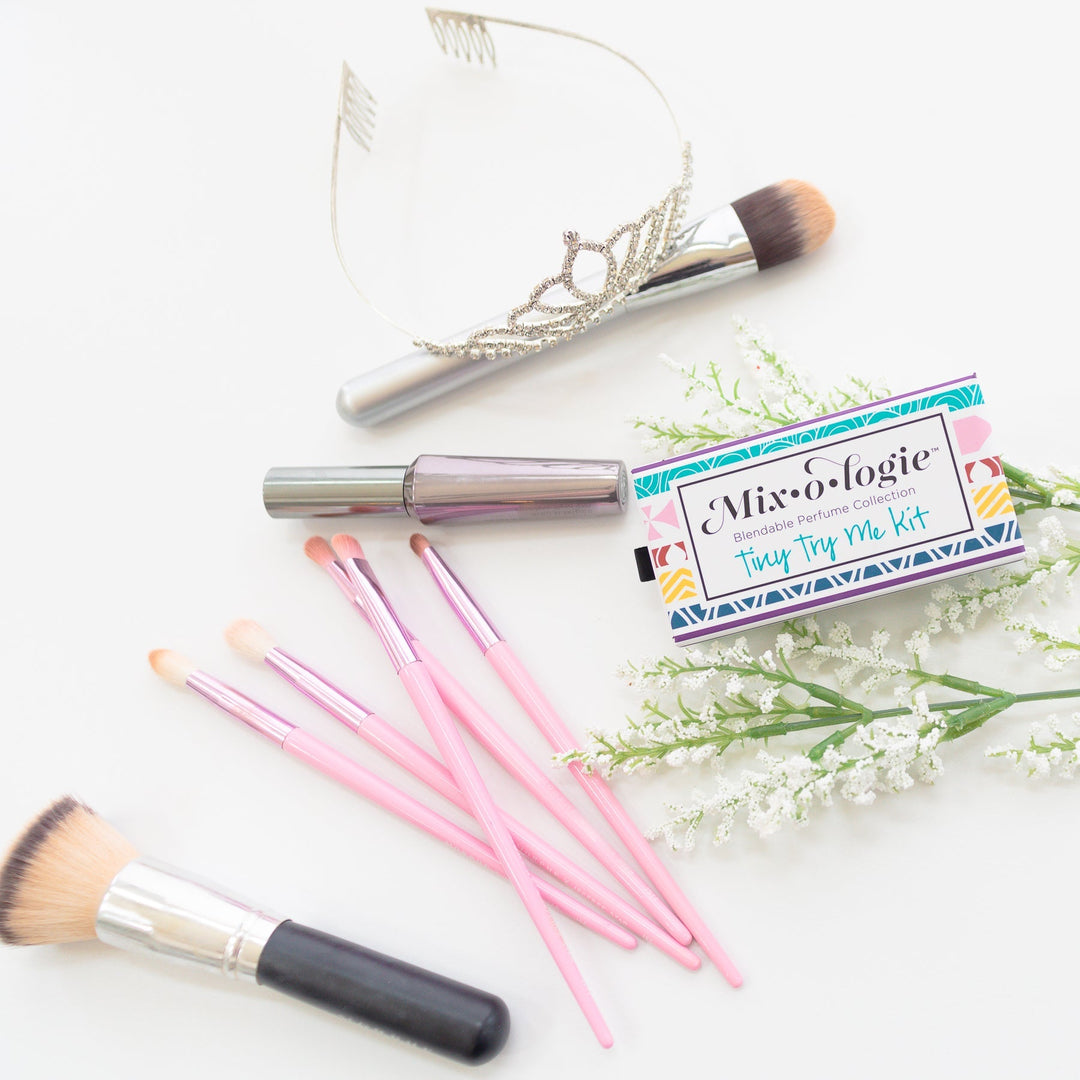 Mixologie Tiny Try Me Kit closed slide box styled flat lay with silver tiara, iridescent mascara tube, pink makeup brushes, and white wildflowers on white background