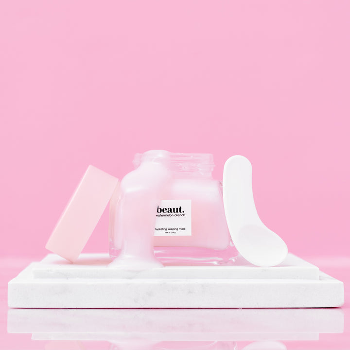 Beaut Watermelon Drench Hydrating Sleeping Mask 50g open glass jar with lid and spatula displayed on white marble tray against pink background